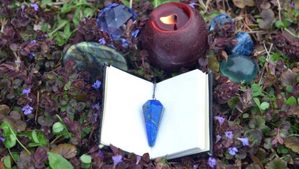 Lapis lazuli pendulum on an open journal, surrounded by a red candle and healing crystals in a garden setting, symbolizing spiritual clarity and inner truth.