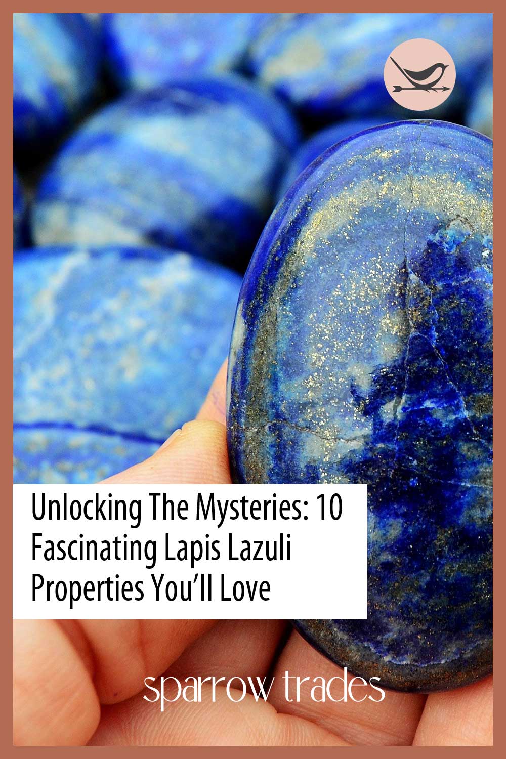 Hand holding a polished oval lapis lazuli stone with gold flecks and deep celestial blue tones, surrounded by similar stones in the background.
