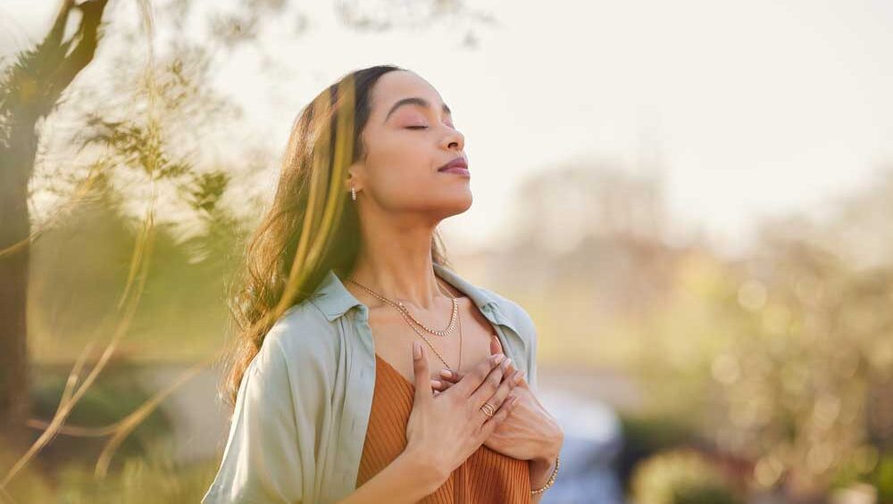Woman standing outside with eyes closed, hands over heart, embracing emotional healing and inner peace.