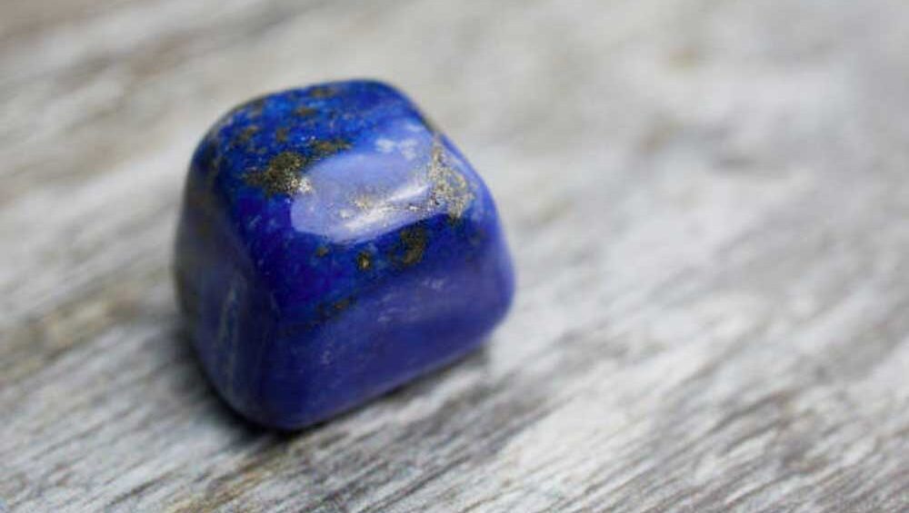 Tumbled lapis lazuli gemstone with deep blue hues and gold flecks on a weathered wood surface