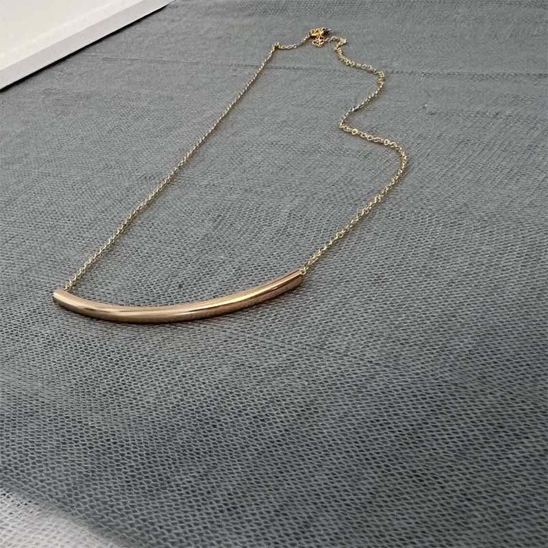 Minimalist curved tube bar necklace in gold filled metal laid out on a soft gray textured fabric background.