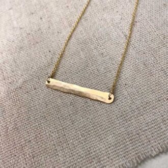 Hand-hammered gold bar necklace with delicate chain, displayed on textured neutral fabric.