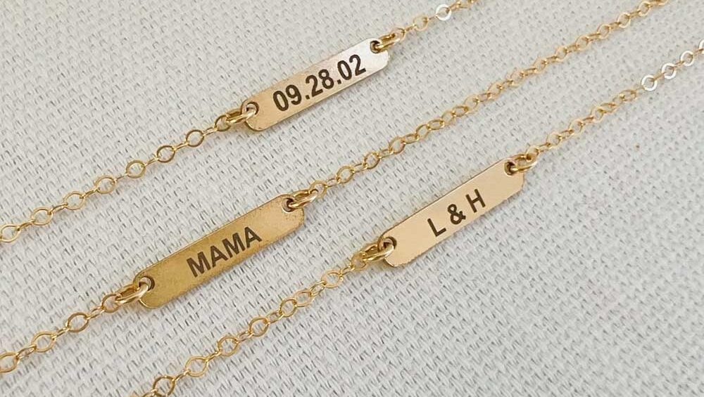 Gold engraved bar necklaces with custom inscriptions including a date, the word "MAMA," and initials "L & H."
