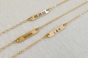 Gold engraved bar necklaces with custom inscriptions including a date, the word "MAMA," and initials "L & H."