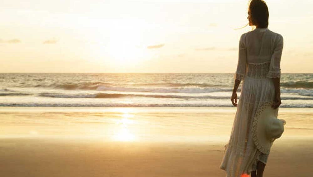 Woman standing alone on the beach at sunset, holding a hat, looking toward the horizon in quiet reflection.