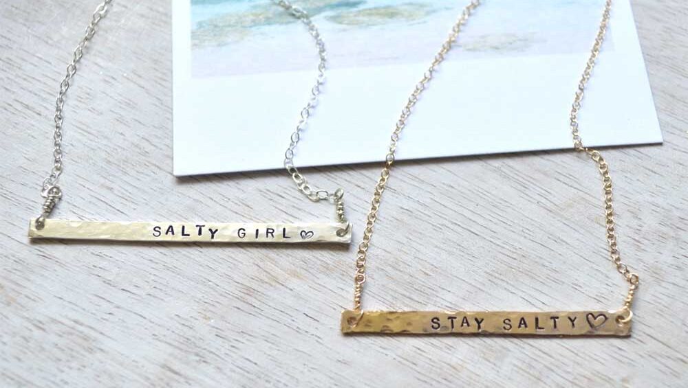 Gold and silver bar necklaces engraved with the phrases “STAY SALTY” and “SALTY GIRL” displayed side by side.