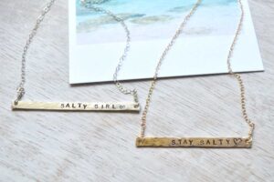 Gold and silver bar necklaces engraved with the phrases “STAY SALTY” and “SALTY GIRL” displayed side by side.