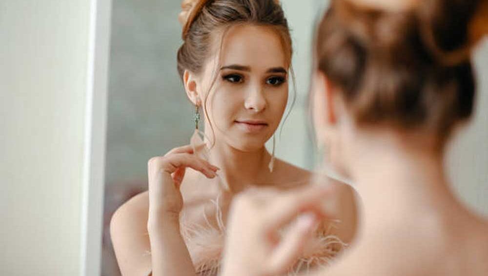 Woman in soft lighting looking at herself in the mirror while wearing delicate earrings.