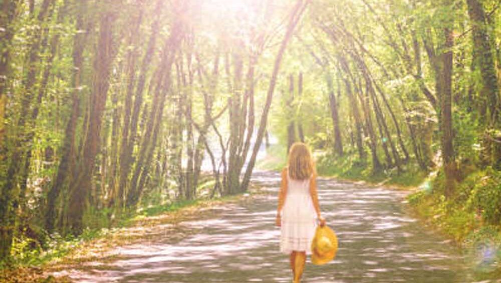 Woman in a white dress walking alone down a sunlit forest path, holding a yellow sunhat.