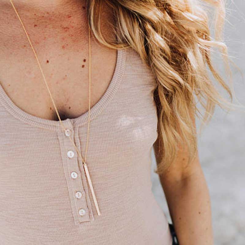 Close-up of a woman wearing a long hammered bar necklace in gold over a buttoned ribbed tank top, with soft blonde waves cascading over her shoulder.