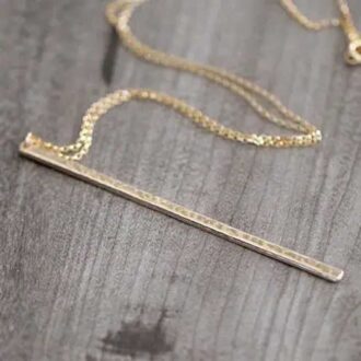 Hammered long bar necklace in gold laid flat on a dark wood surface, showing the bar’s shine and textured detail.