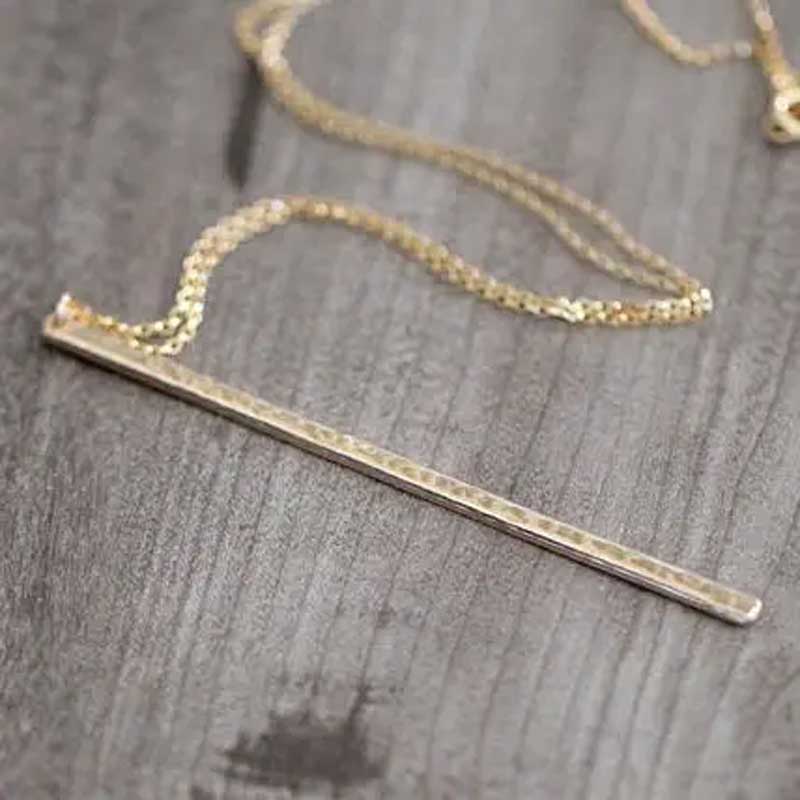 Hammered long bar necklace in gold laid flat on a dark wood surface, showing the bar’s shine and textured detail.