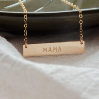 Close-up of a hand-stamped gold bar necklace reading “MAMA,” hanging on a delicate gold chain against a neutral background.