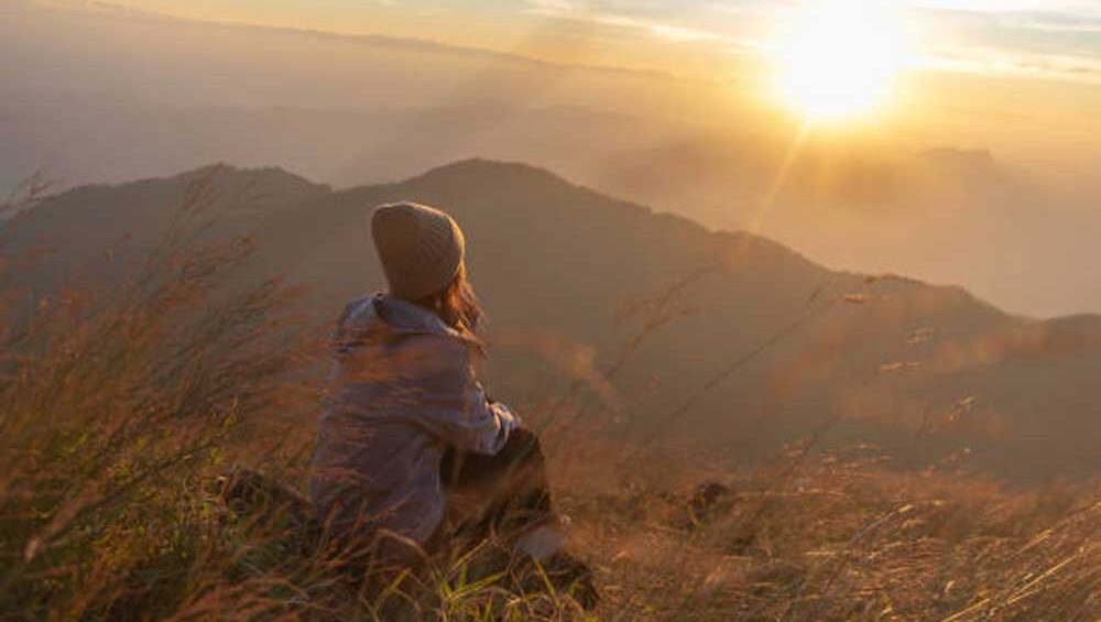 Woman sitting in tall grass, gazing at the sunrise over mountain peaks