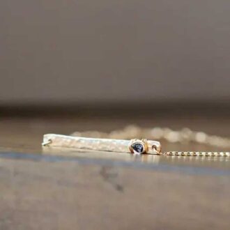 Close-up of a hammered gold bar necklace with a raw black diamond wrapped in gold wire, resting on a wooden surface.