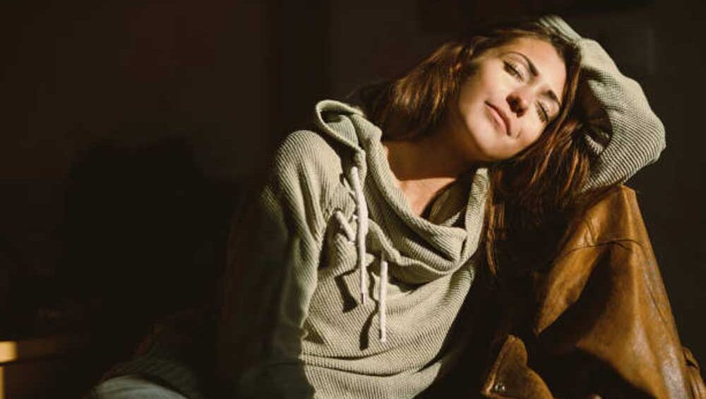 Woman in soft light wearing a green hoodie, leaning into herself with a grounded, emotionally clear expression.
