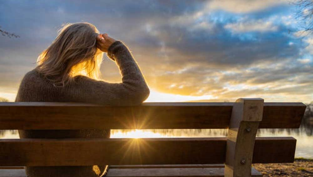 A woman sitting alone on a bench at sunset, resting her head in thought during a moment of quiet reflection.