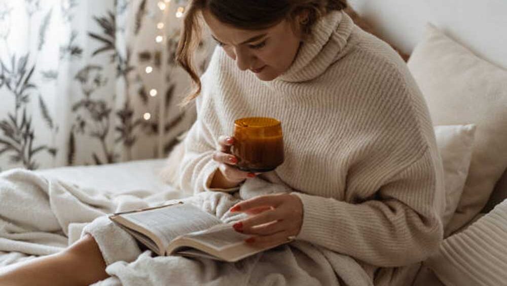 Woman in cozy sweater reading a book with a warm drink, reflecting in a peaceful, soft-lit room.