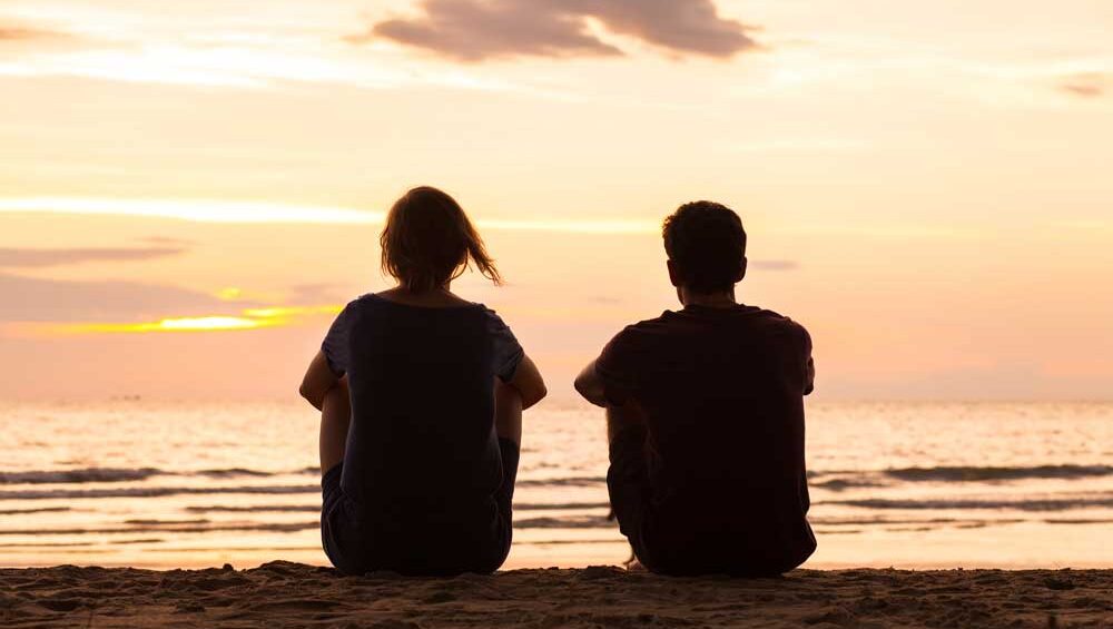 Couple sitting side by side on the beach at sunset, symbolizing emotional distance and quiet connection.