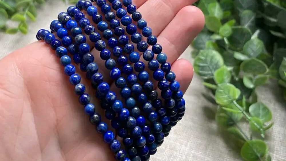 Hand holding several strands of round lapis lazuli bead bracelets, showcasing deep blue hues with gold flecks against a natural leafy background.