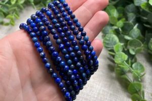 Hand holding several strands of round lapis lazuli bead bracelets, showcasing deep blue hues with gold flecks against a natural leafy background.