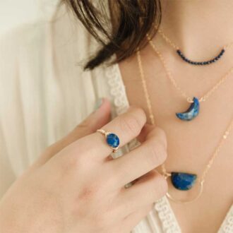 Hand wearing a gold-filled oval lapis lazuli ring, styled with layered blue gemstone necklaces.
