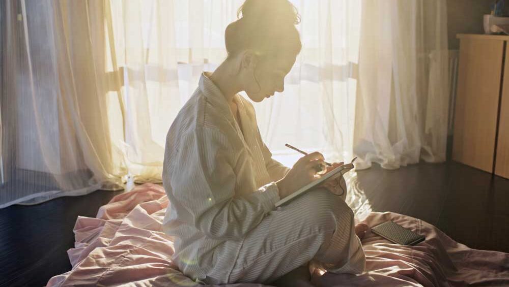 Woman sitting on a bed writing in her journal in soft morning light
