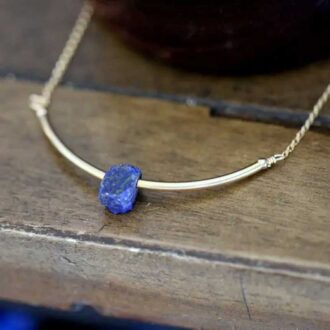Close-up of the Valkyrie Lapis Lazuli Necklace featuring a rough-cut lapis nugget set in a curved 14k gold-filled bar on a wood surface.