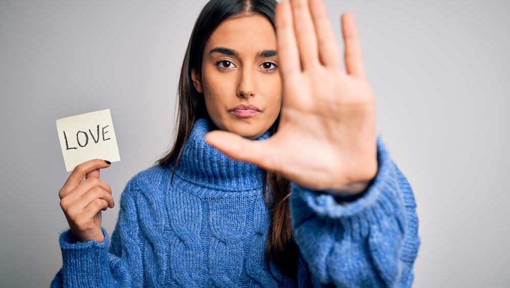Woman holding a note that says “LOVE” while raising her hand in a stop gesture, symbolizing emotional avoidance.