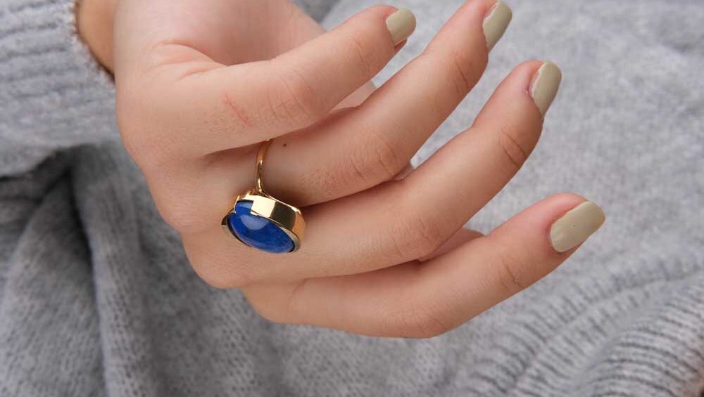 Close-up of a woman's hand wearing a gold lapis lazuli ring, styled with neutral nail polish and a soft gray sweater.