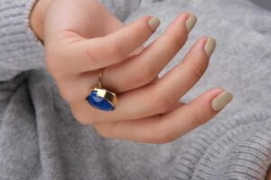 Close-up of a woman's hand wearing a gold lapis lazuli ring, styled with neutral nail polish and a soft gray sweater.