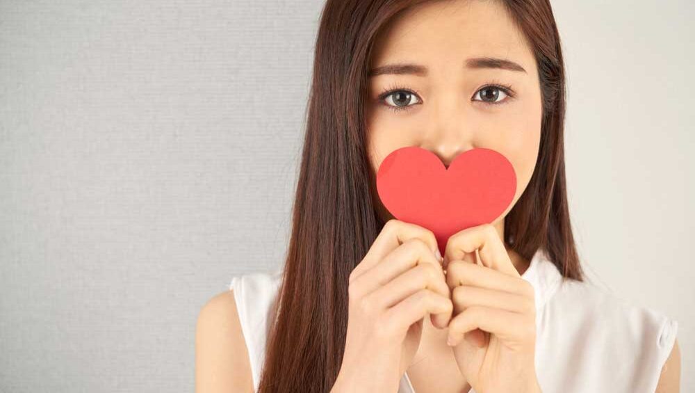 A sad woman holds a red paper heart in front of her mouth, symbolizing unspoken love and emotional silence in a strained mother-daughter relationship.