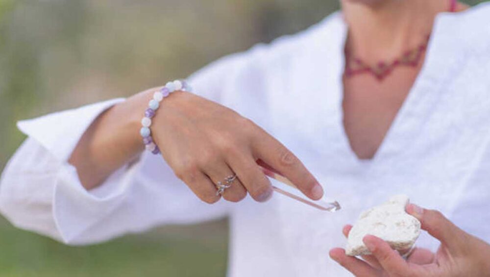 Woman using tweezers to select a gemstone from a cluster while wearing a pastel crystal bracelet, symbolizing intentional healing jewelry practice.
