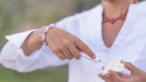 Woman using tweezers to select a gemstone from a cluster while wearing a pastel crystal bracelet, symbolizing intentional healing jewelry practice.
