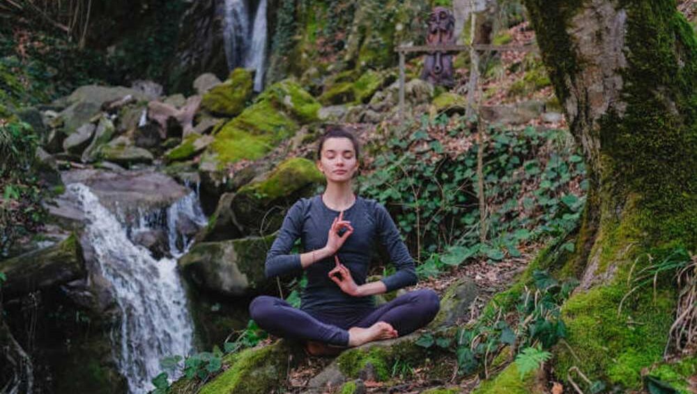 Woman sitting cross-legged in meditation pose near a forest waterfall, practicing mindfulness in nature.