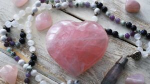 Heart-shaped rose quartz crystal surrounded by a beaded mala necklace with moonstone, amethyst, and black tourmaline on rustic wood background