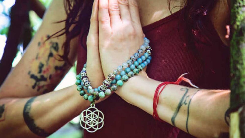 Hands in prayer pose holding Reiki healing jewelry with turquoise beads and sacred geometry pendant in nature