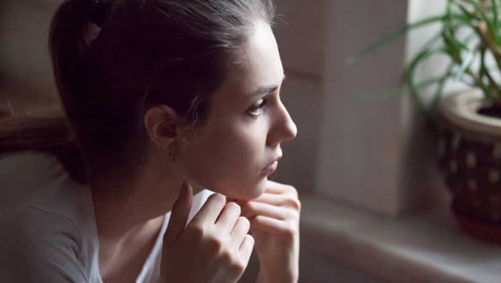 Young woman gazing out the window, lost in thought, symbolizing rumination and overthinking.