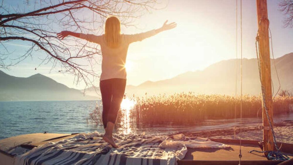 Woman standing barefoot on a dock with arms outstretched toward sunrise over a peaceful lake, symbolizing the freedom of disconnecting with love.
