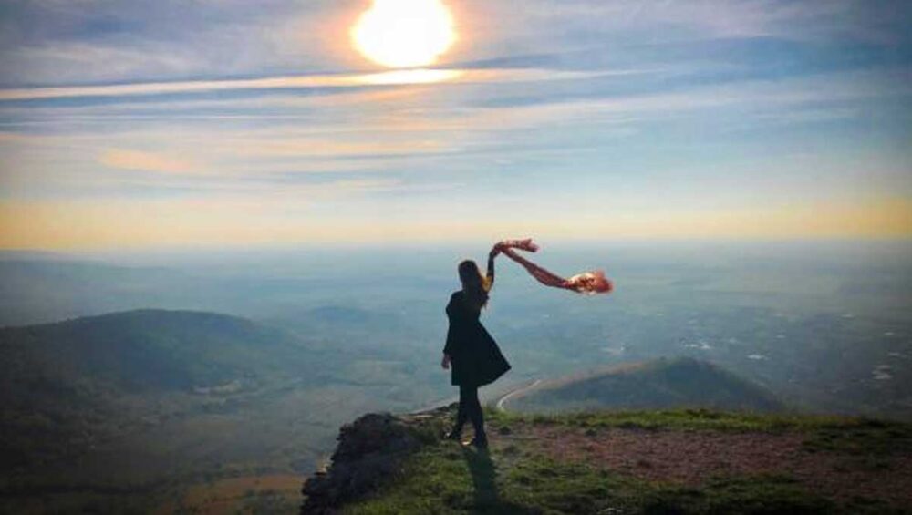 Woman standing on a cliff at sunrise, scarf blowing in the wind, symbolizing letting go and finding freedom.