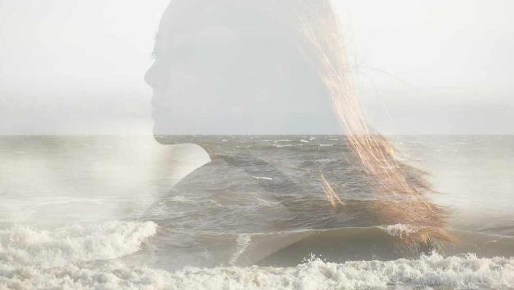 Double exposure of a woman's silhouette blending with the ocean waves, symbolizing emotional erasure and inner disconnection.