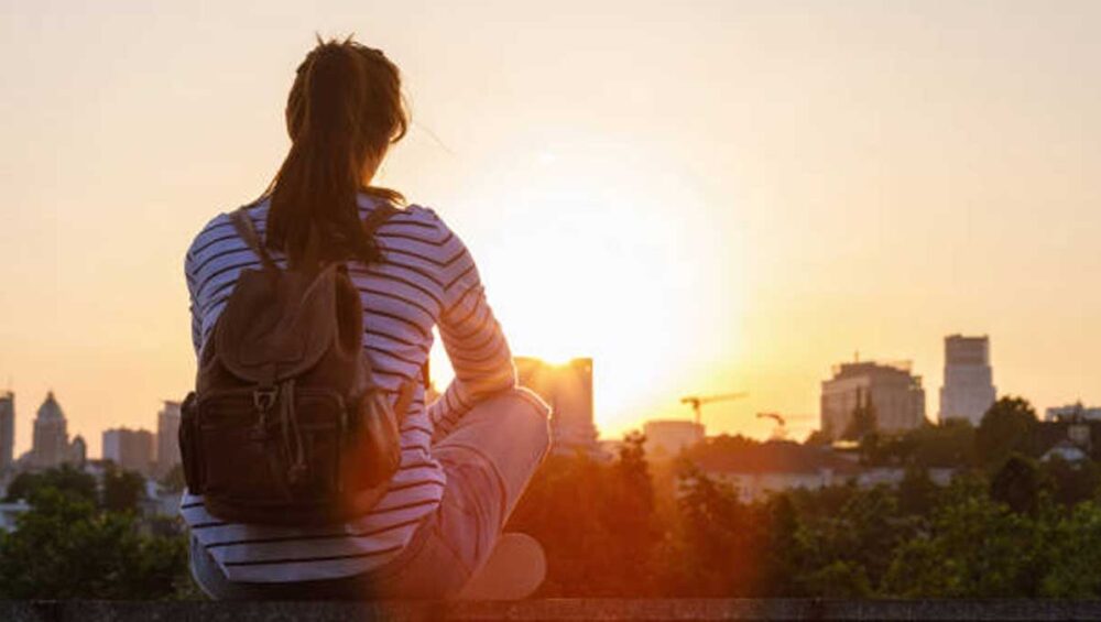 Woman with backpack sitting quietly on a rooftop at sunset, looking over the city skyline