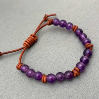 Adjustable amethyst bead bracelet on rustic brown leather with a sliding-knot clasp on a soft gray background.