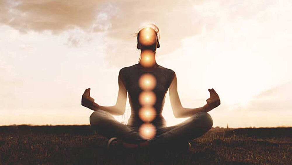 Person meditating with glowing chakras aligned along the spine at sunset