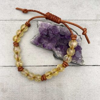 Adjustable citrine bead bracelet on rustic brown leather with a sliding-knot clasp beside an amethyst cluster on a gray wooden surface.