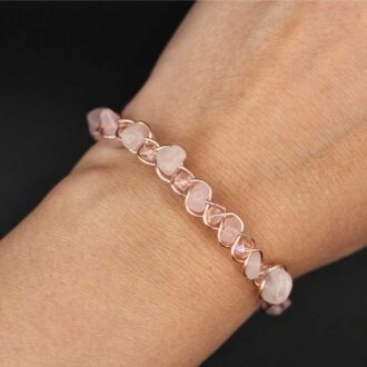 Hand wearing a rose quartz chip bangle braided with rose-gold wire and tiny faceted glass accents, finished with a hook clasp on a black background.