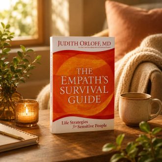 The Empath's Survival Guide book displayed on a wooden table beside a ceramic mug, candle, notebook, and greenery in a warmly lit room.