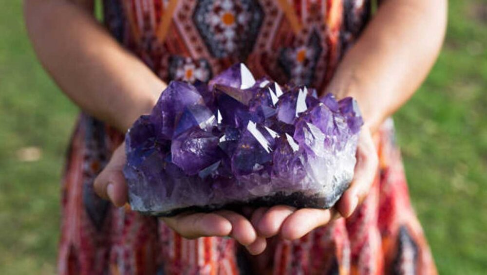 Hands holding a large raw amethyst crystal cluster outdoors