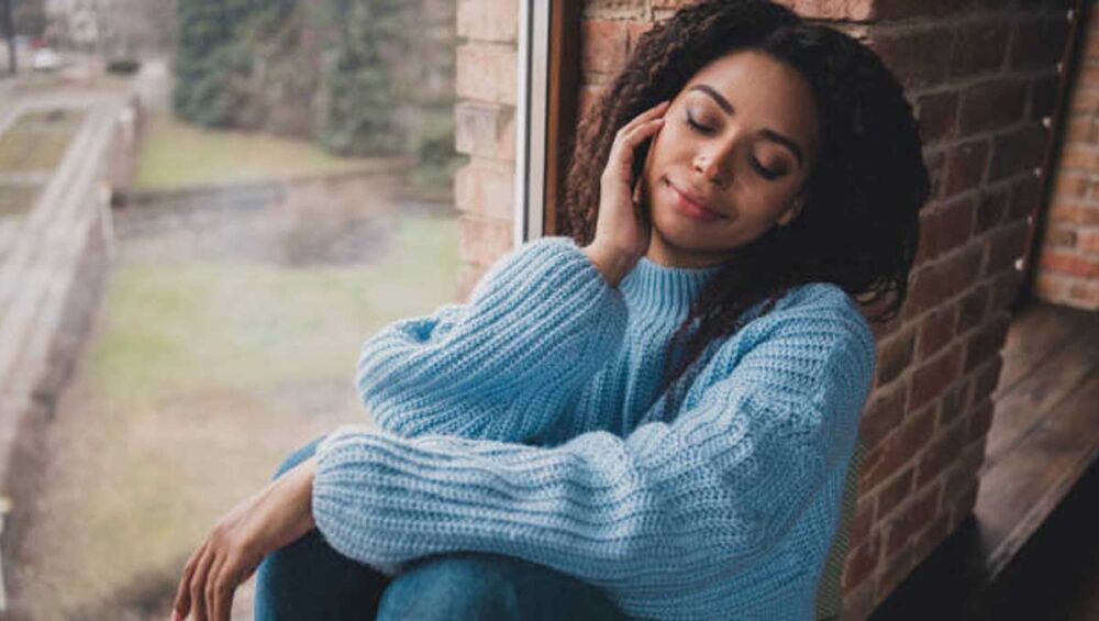 woman sitting by a window in a blue sweater with eyes closed, reflecting calmly