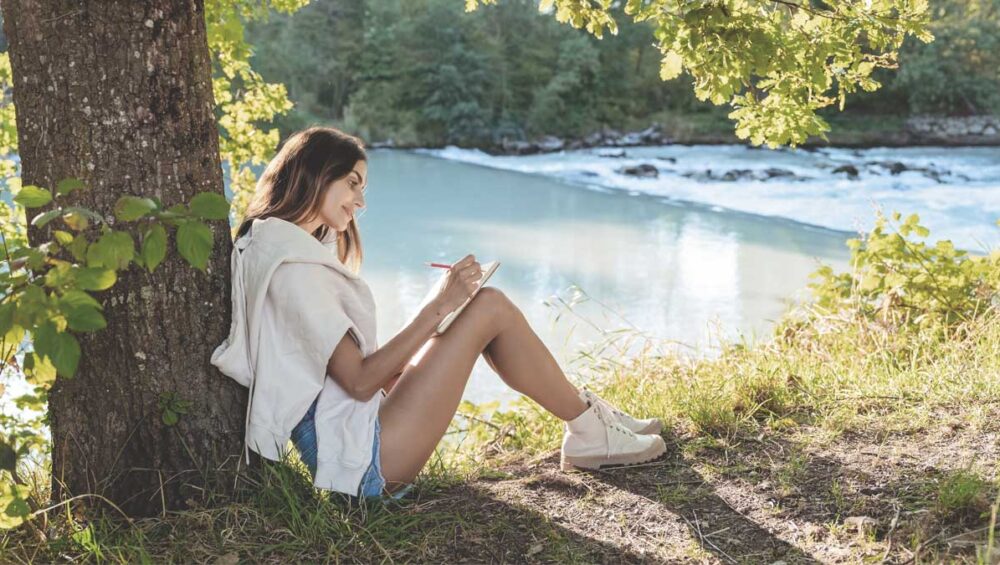 woman journaling by a calm river under sunlight, reflecting and reconnecting with her emotions